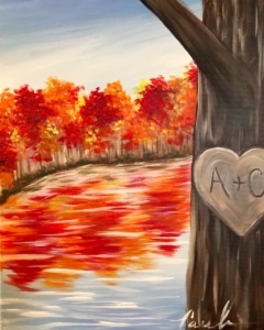 A romantic fall landscape with red and orange trees reflecting on a calm lake and a carved heart on a tree trunk – Wine & Canvas Toledo painting “Fall Lake”