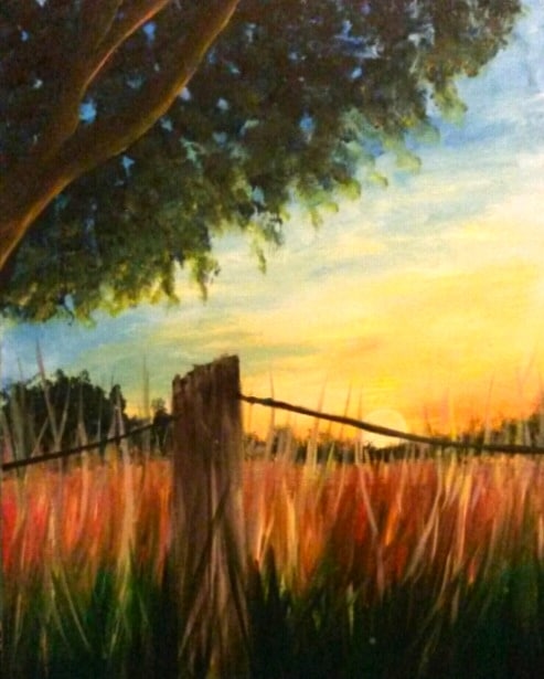 A rustic wooden fence post with barbed wire stretches across a glowing sunset field, surrounded by tall golden grasses and framed by a leafy tree against a warm orange and blue evening sky.