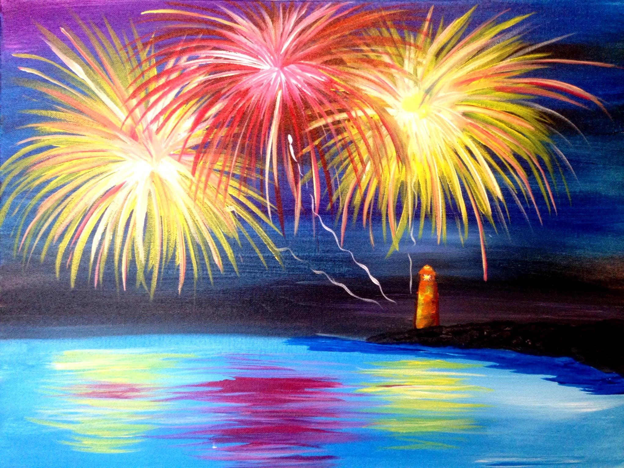 Colorful fireworks exploding over calm beach water beside a glowing lighthouse at night with bright reflections across the ocean.