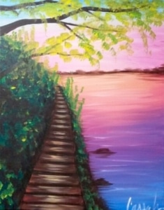 A narrow wooden boardwalk winds through lush green foliage along the edge of a calm lake, beneath overhanging tree branches and a soft pink and purple sunset sky.